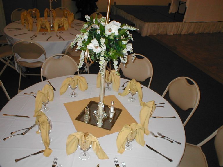 A round banquet table set for a formal event, featuring cream-colored napkins folded in glasses, a floral centerpiece with white flowers, and neatly arranged silverware around each place setting.