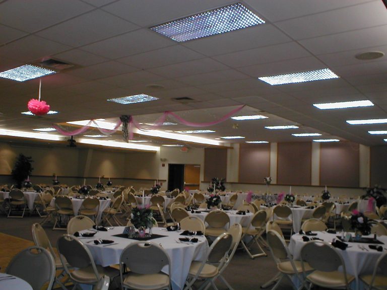 A decorated banquet hall with round tables set for a formal event. Each table has a white tablecloth, black napkins, and floral centerpieces. Pink fabric and decorations hang from the ceiling, and chairs are arranged around the tables.