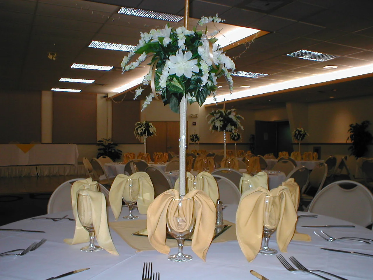 A banquet hall is set up for an event with round tables covered in white tablecloths. Each table features a tall floral centerpiece with white flowers and greenery, and is surrounded by chairs with cream-colored napkins folded in wine glasses.